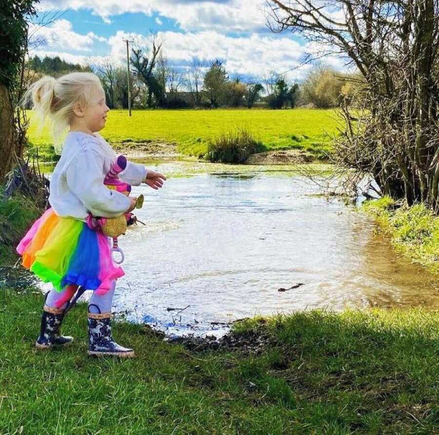 Little girl in rainbow tutu and rain boots runs around outside near a pond