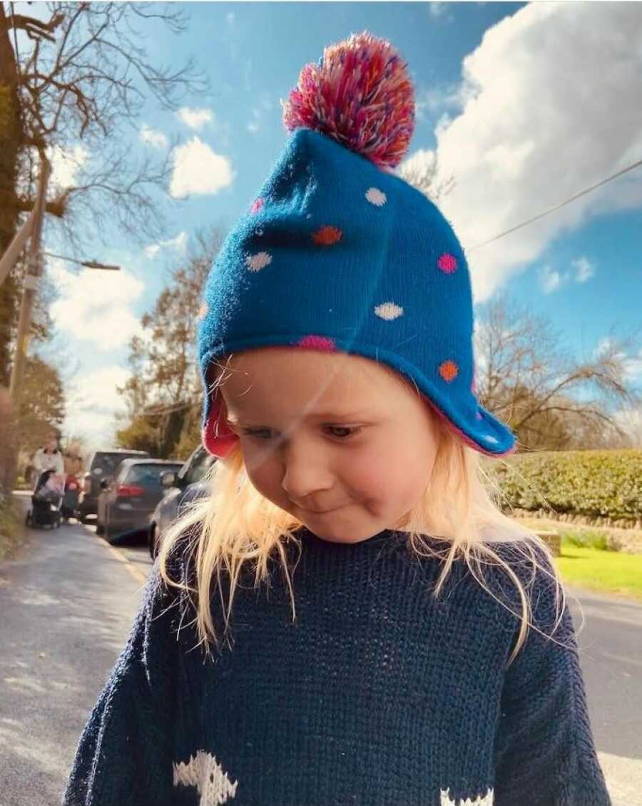 Little girl looks down towards the ground while on a walk, showing off her pink and blue beanie