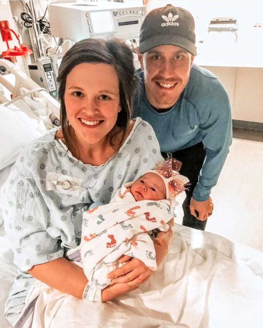 Mom holding newborn baby girl at the hospital with her husband leaning from behind 