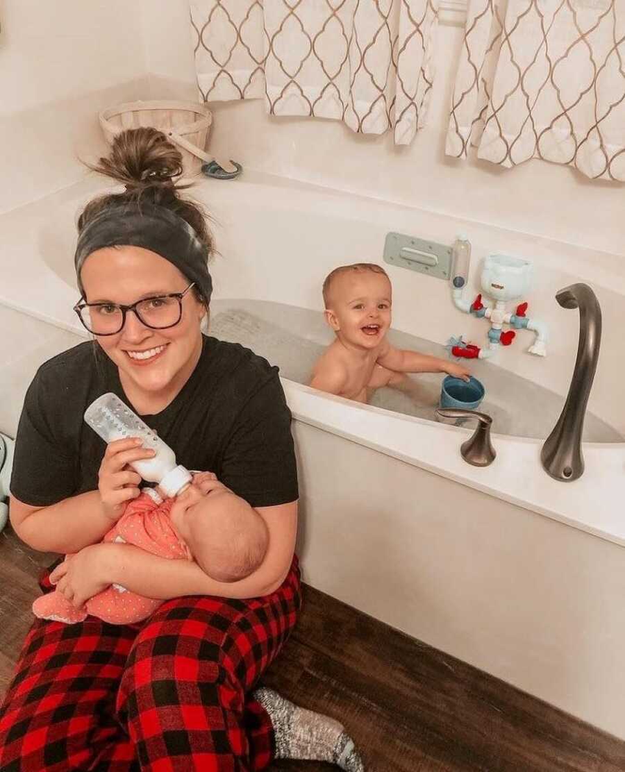Mom bottle feeding her newborn baby girl while toddler son takes a bath 