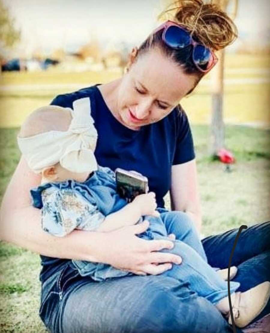 Gestational carrier sitting on the grass holding baby girl with a big white bow on her head 