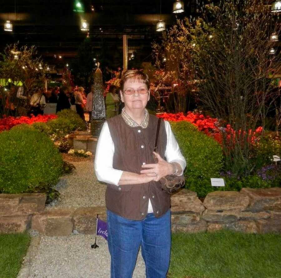 Woman stands in front of floral scenery in a brown vest and jeans