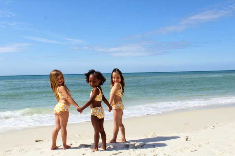 Foster daughters in matching yellow bathing suits smile for a photo while holding hands on the beach