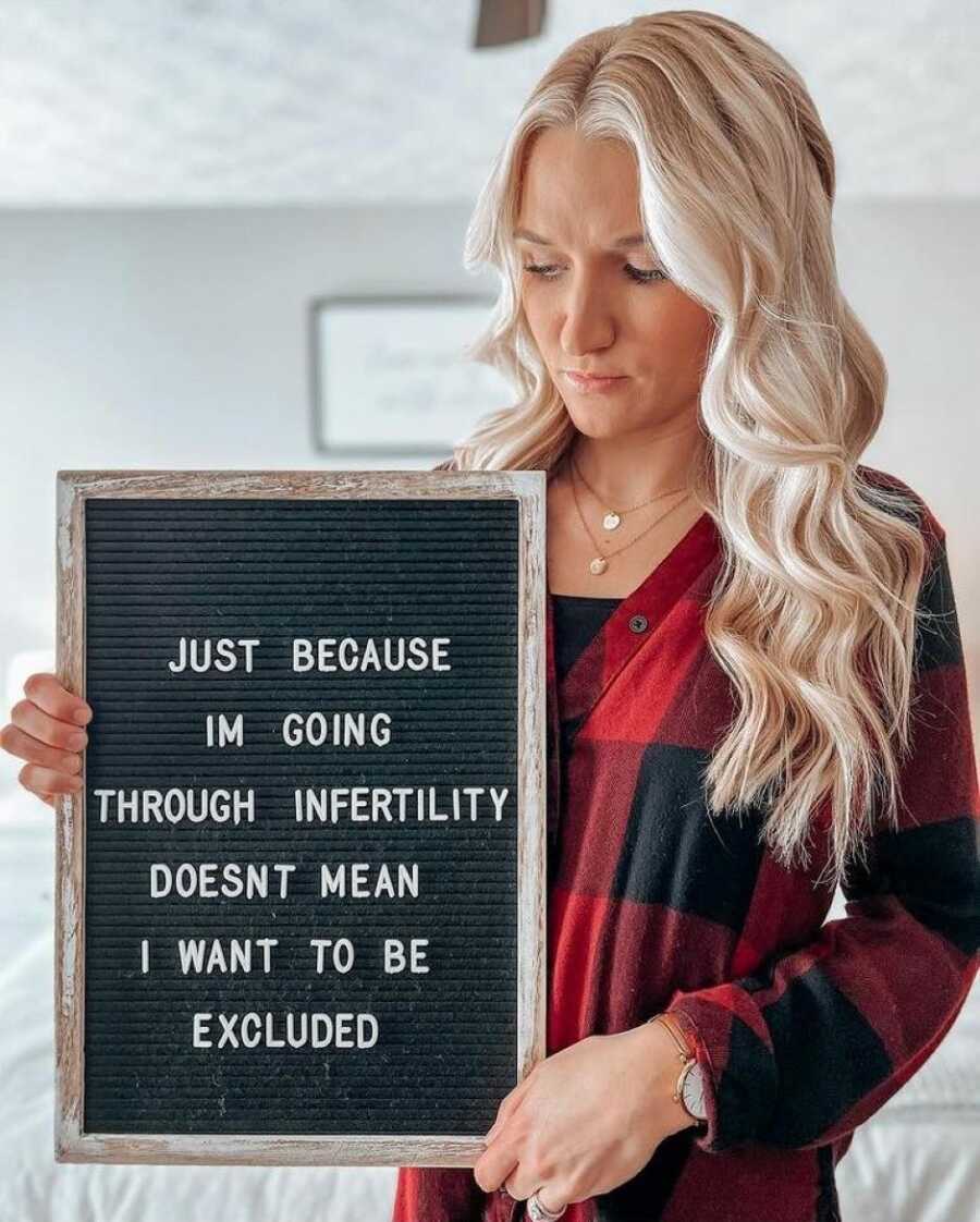 Woman battling infertility looks somber in red and black shirt while holding a sign about being excluded from events due to her infertility battle