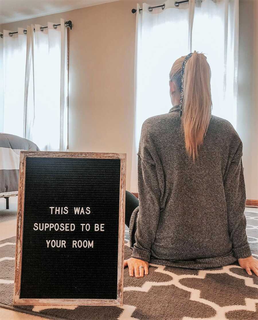 woman battling infertility sitting in a room meant to be the nursery room for her long-wanted baby with a sign on the floor that says "This was supposed to be your room"