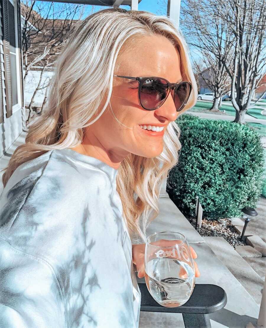 Woman battling infertility sitting on a porch with a glass of water on her hand, wearing sunglasses, and smiling on a sunny day 