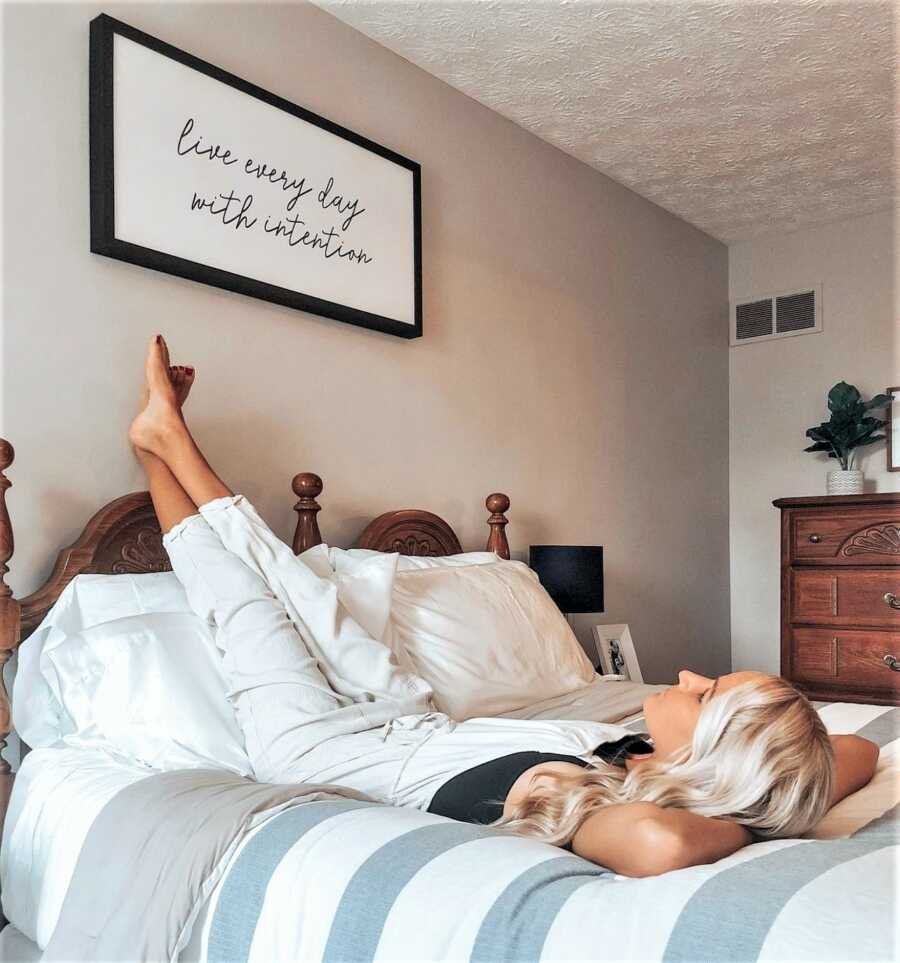 Young woman laying upside-down on a bed with her feet resting on the wall and crossed arms under her head while staring at a sign on the wall saying "LIVE EVERY DAY WITH INTENTION"