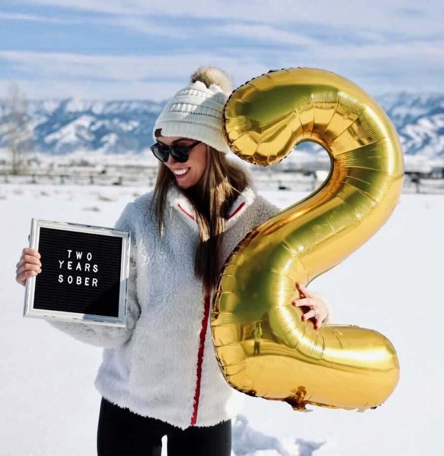Woman celebrates two years sober with a "2" golden balloon and a photoshoot in an all-white winter outfit in the snow