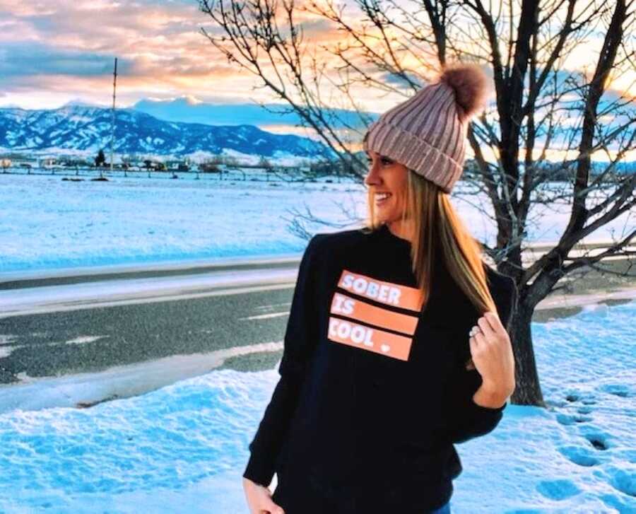 Woman recovering from alcoholism wears a "sober is cool" sweatshirt while enjoying the sunset in the snow