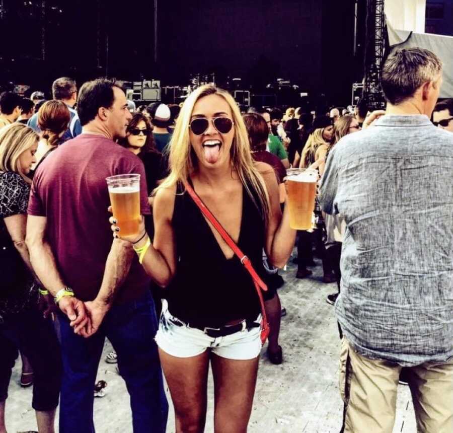 Woman battling alcoholism holds two beers while sticking her tongue out, waiting for a concert to start at a packed venue