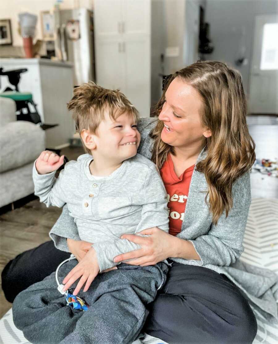 special needs mom holding son while both sitting on the Livingroom floor staring at each other smiling 