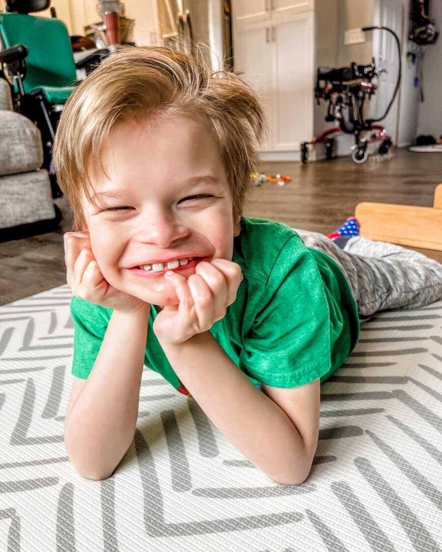 Little boy with rare disease smiles big for a photo while laying on the floor in a green t-shirt