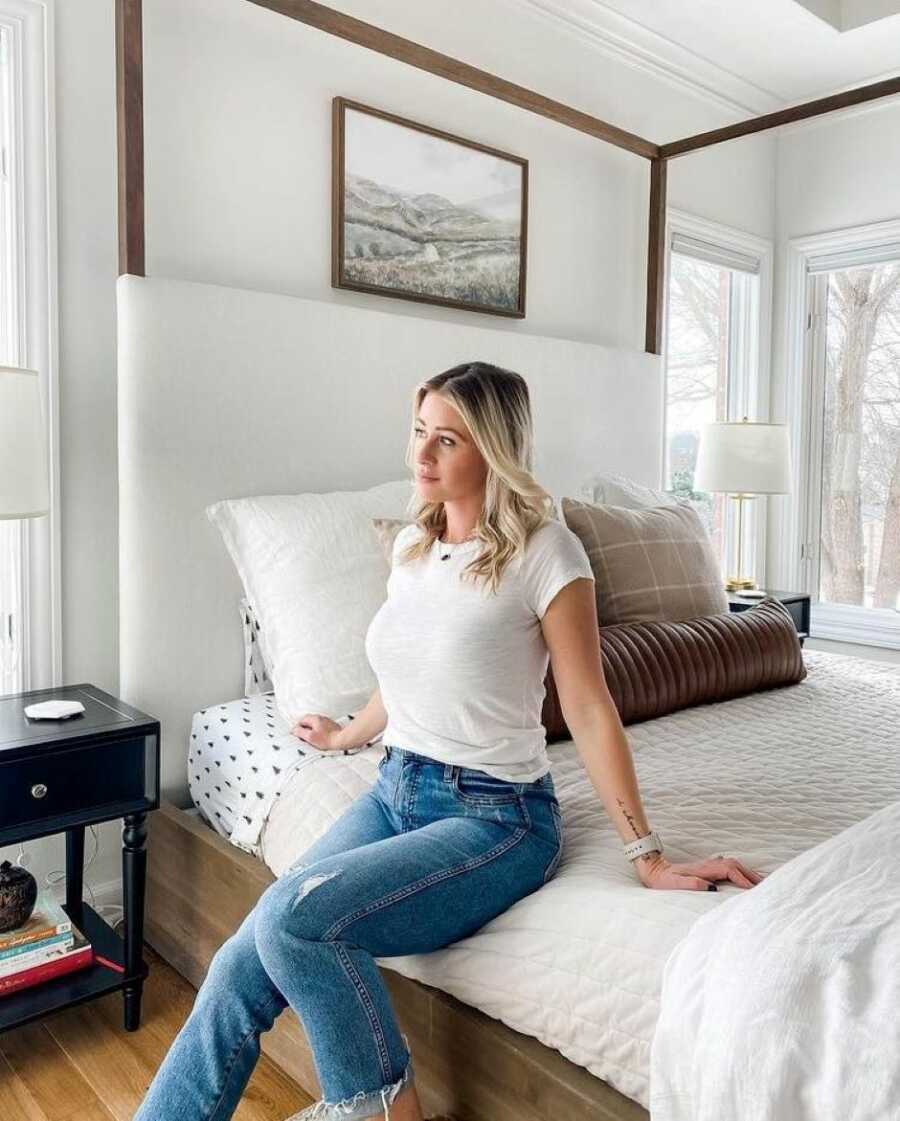 Remarried widow looks reflective while sitting on her bed in a white t-shirt and jeans