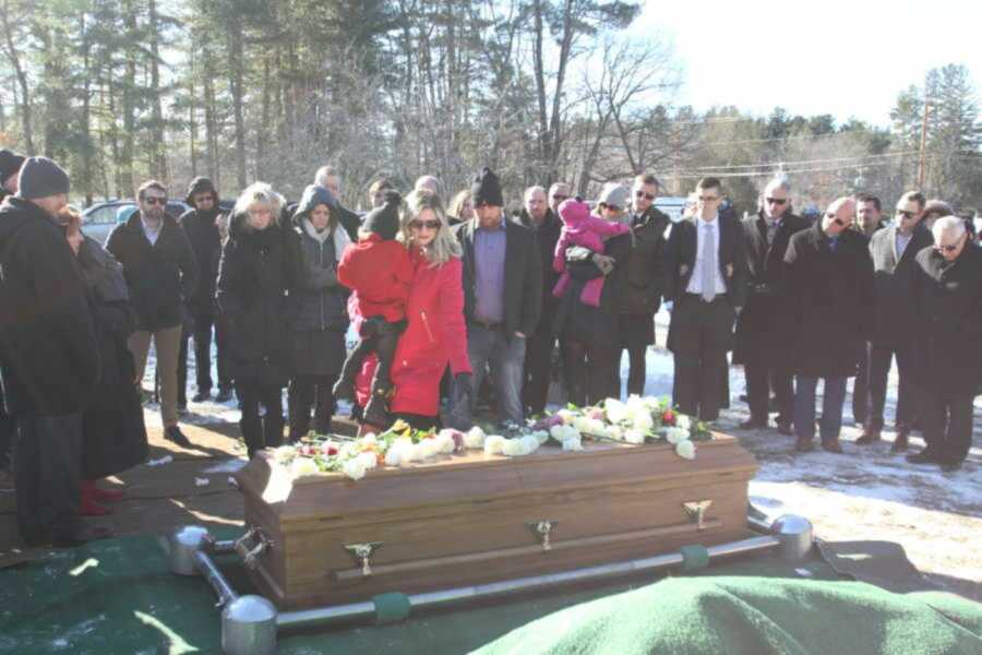 Woman in red coat holds one of her children while putting flowers on her late husband's casket