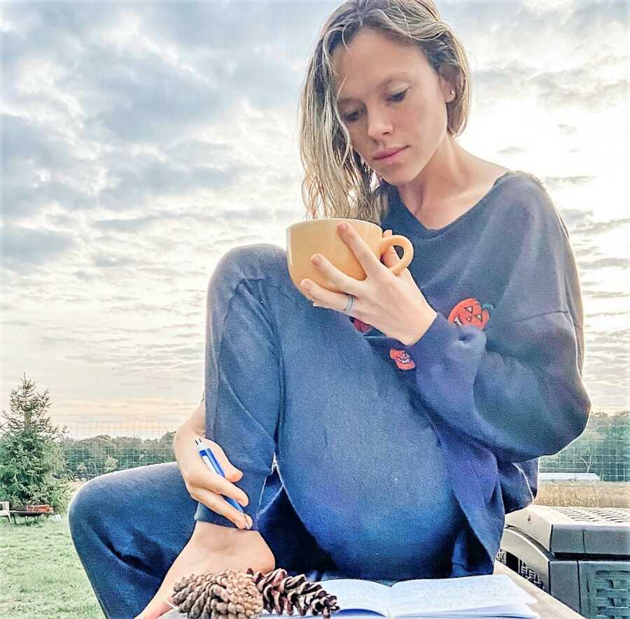 woman sitting outdoors writing on a notebook wearing comfy clothes holding a mug 