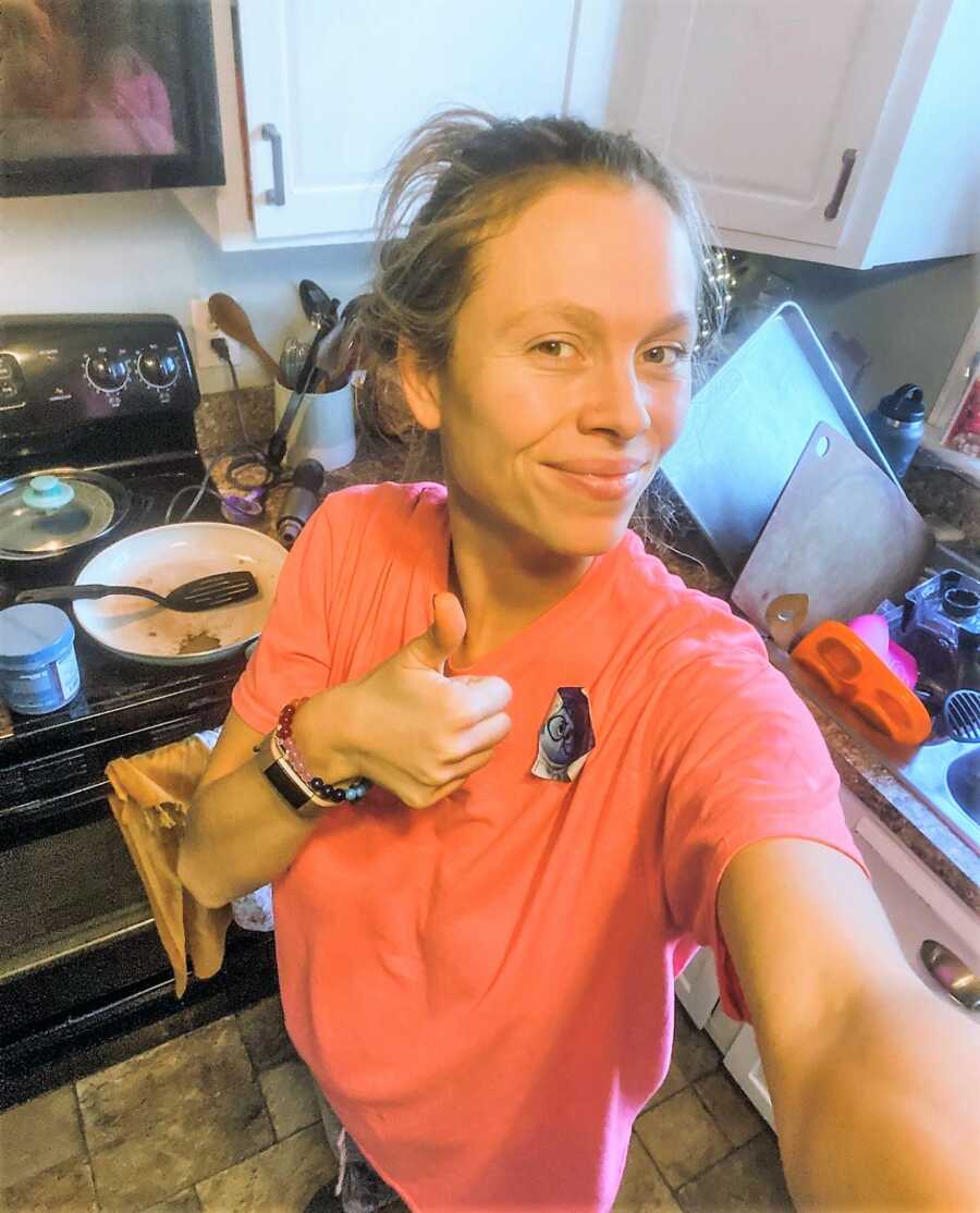 selfie of mom with a pile of dirty dishes in the back and a messy kitchen while smiling with her thumb up