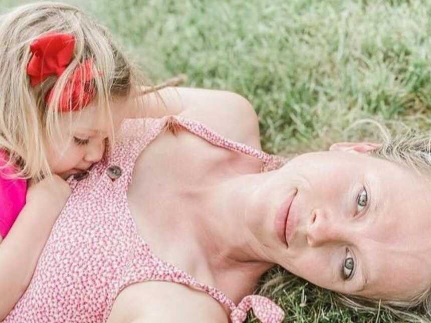 Woman lays in the grass in floral dress while her daughter lays on top of her