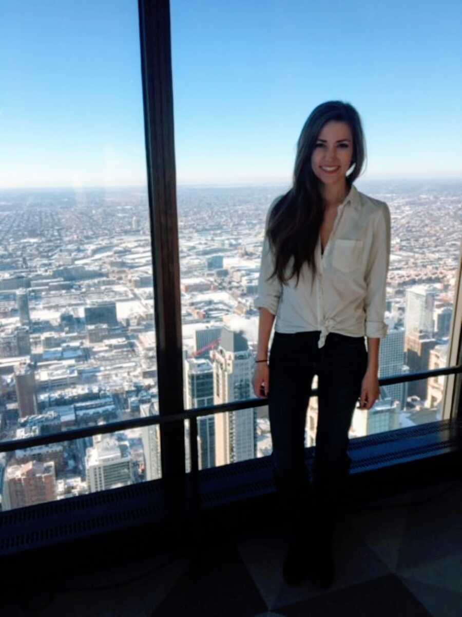 Woman in white button-down shirt and black skinny jeans smiles for a photo in front of a city landscape behind her