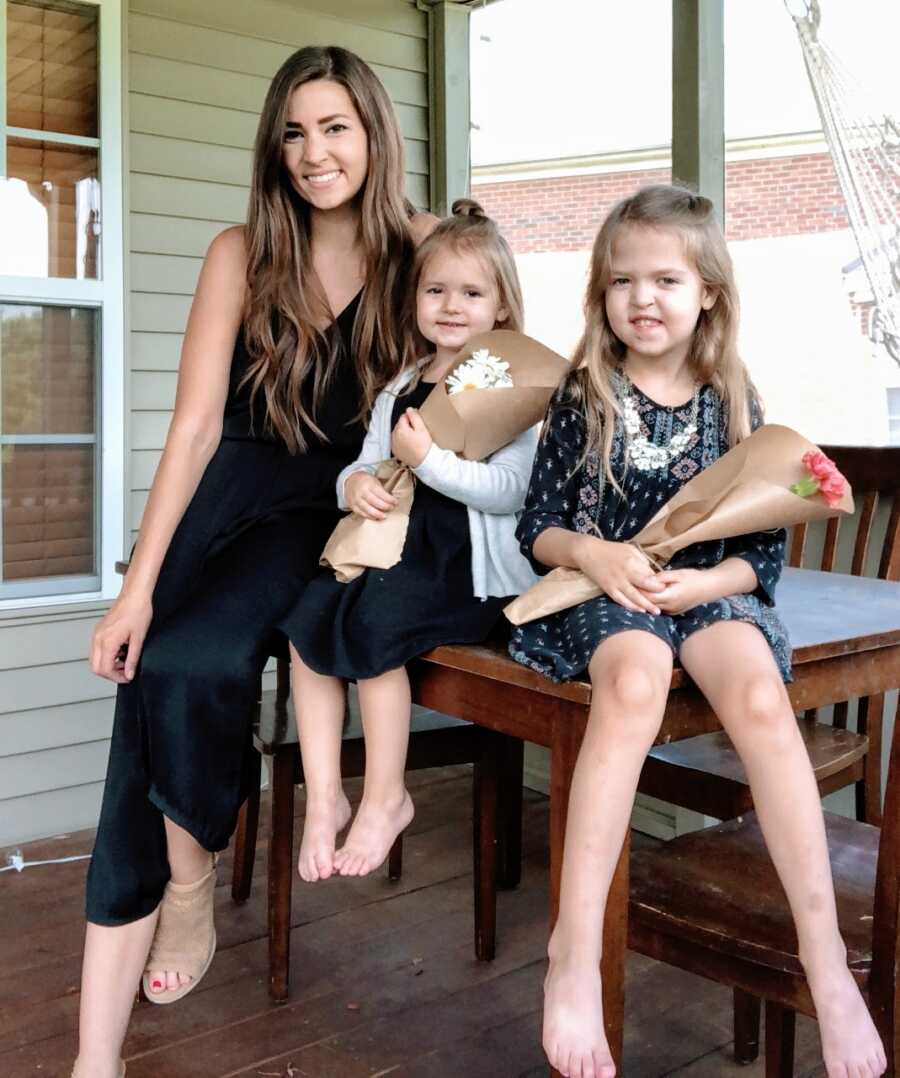 Woman in black dress takes a photo with her daughters, both holding a bouquet of flowers, while sitting on a table on their porch