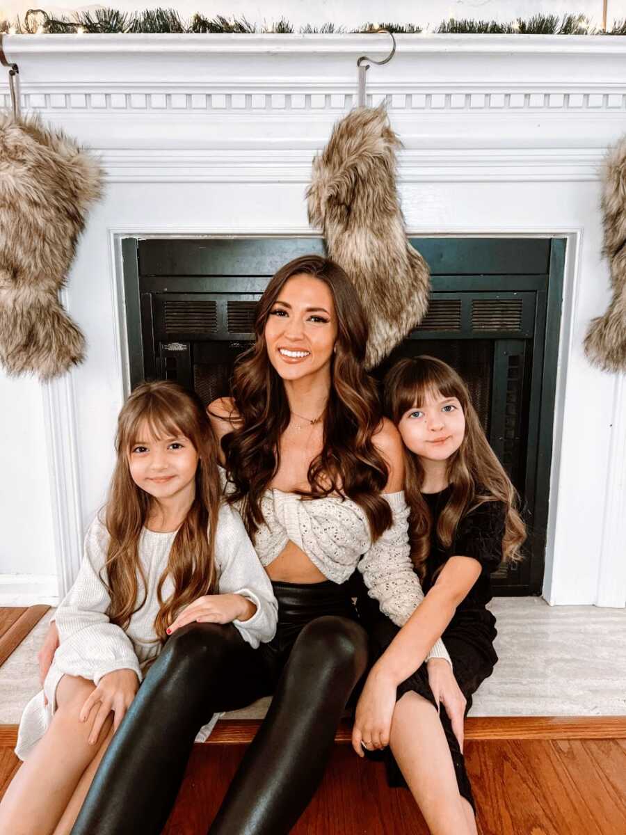 Woman on self-love journey takes a photo with her two daughters while they sit on a white fireplace