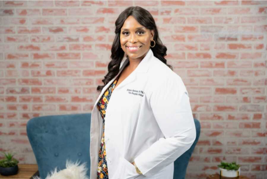 African American nurse wearing a white lab coat and smiling with her body turned to the side and a brick wall in the background