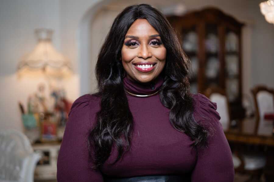 Mid shot of African American woman wearing a long sleeve turtle neck shirt smiling with the Livingroom blurred in the background