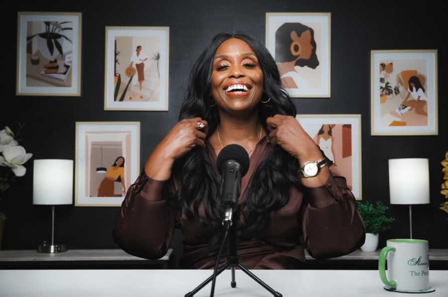 African American woman sitting in front of a microphone while smiling and fixing her shirt's necklace with painted art of African American women in the back