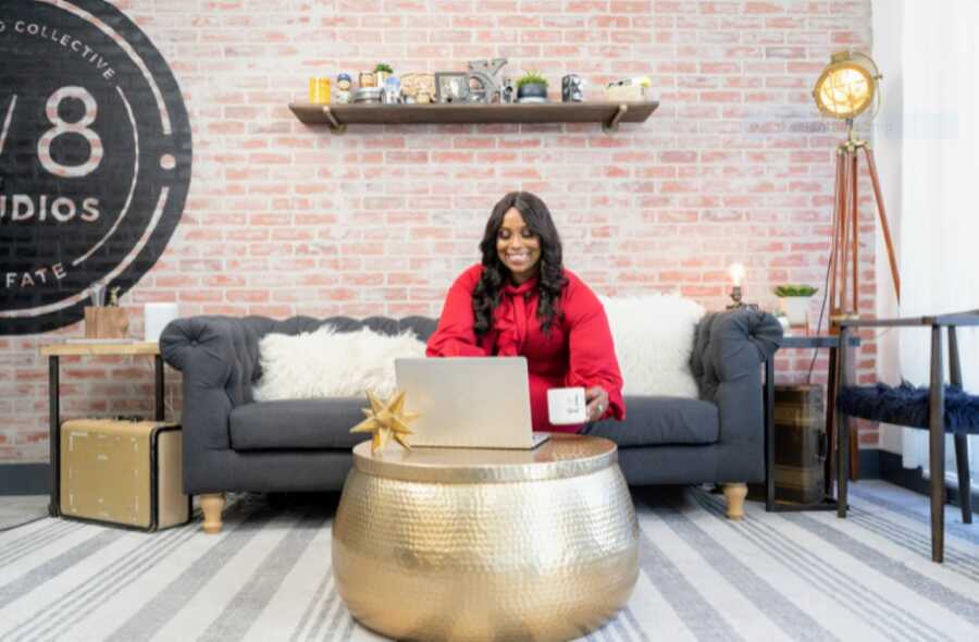 African American woman sitting on a couch at a studio while smiling at the computer and holding a mug