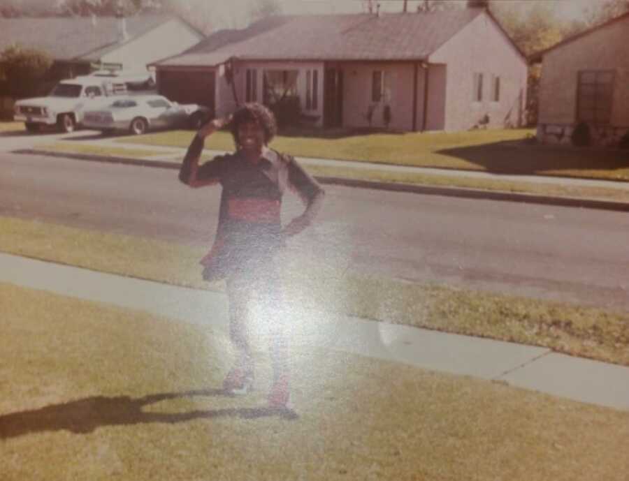 picture of a young African American woman standing on the street outside of her house waving and smiling