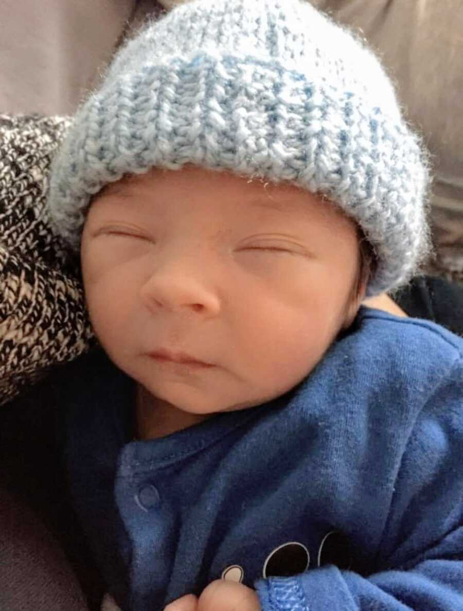 Newborn boy sleeps in his mom's arms while wearing a blue crocheted hat and a blue sweater