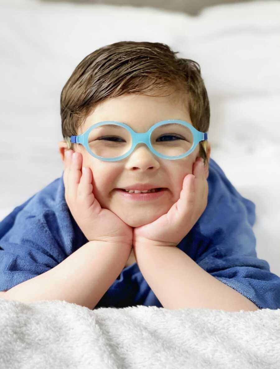 Little boy with rare hearing loss disorder smiles big for a photo in a blue shirt and bright blue glasses