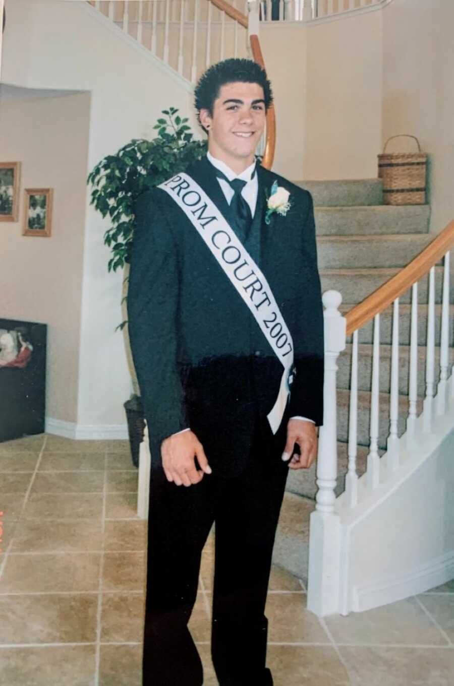 High school student smiles for a photo in a suit while wearing a sash that says "Prom court 2007"