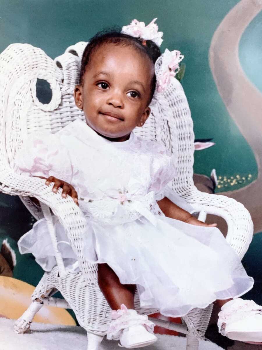 Little girl sits in white whicker chair while wearing a white dress with white bows in her hair