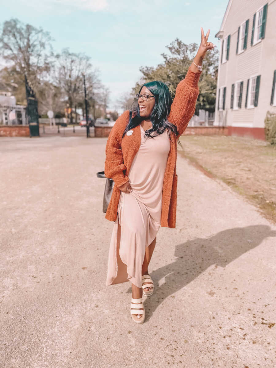 Woman looks happy and blissful while walking in front of her house in a tan dress and orange cardigan