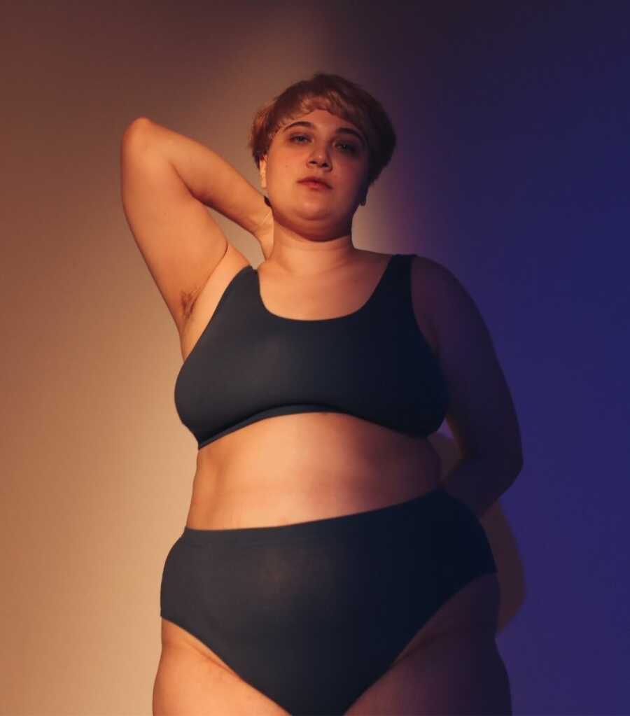 Woman stands in black sports bra and underwear with one arm raised and bent toward her neck, standing in front of a brown and blue shaded backdrop.