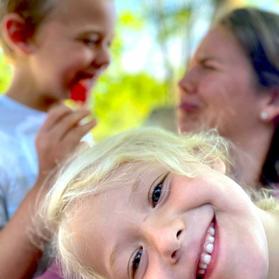 mother with her two kids, one has a big smile close to the camera, the other is with her mom in the back making silly faces