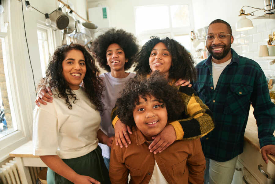 Black and Pakistani mixed race family gather with one another in the kitchen, all are smiling widely