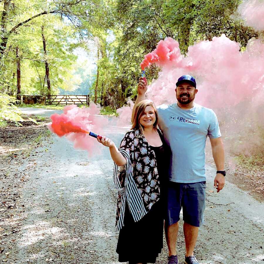 husband and wife pose together for their gender reveal, holding smoke cannons with pink smoke