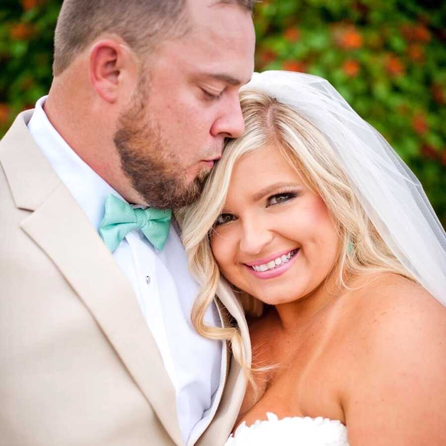 husband kisses his wife's head on their wedding day, she smiles widely