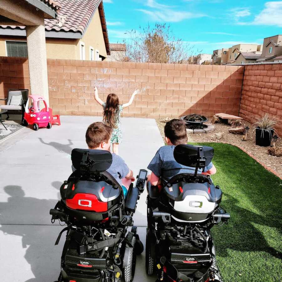 two boys with Duchenne sit in their motor chairs while their sister is playing outside