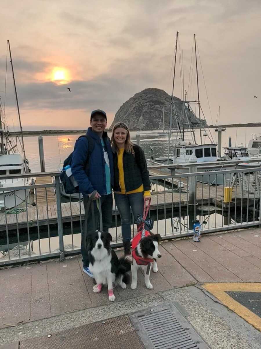 Couple and border collies stand in front of boats on dock at sunset.