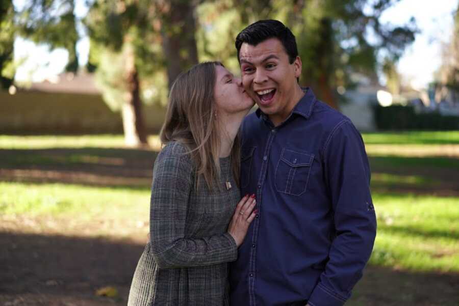 Wife kisses husband on the cheek while he makes an excited face.