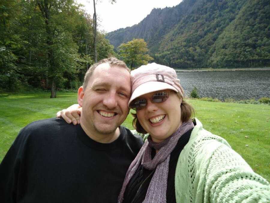 Couple takes picture together with mountains, lake, and pine trees behind them. 