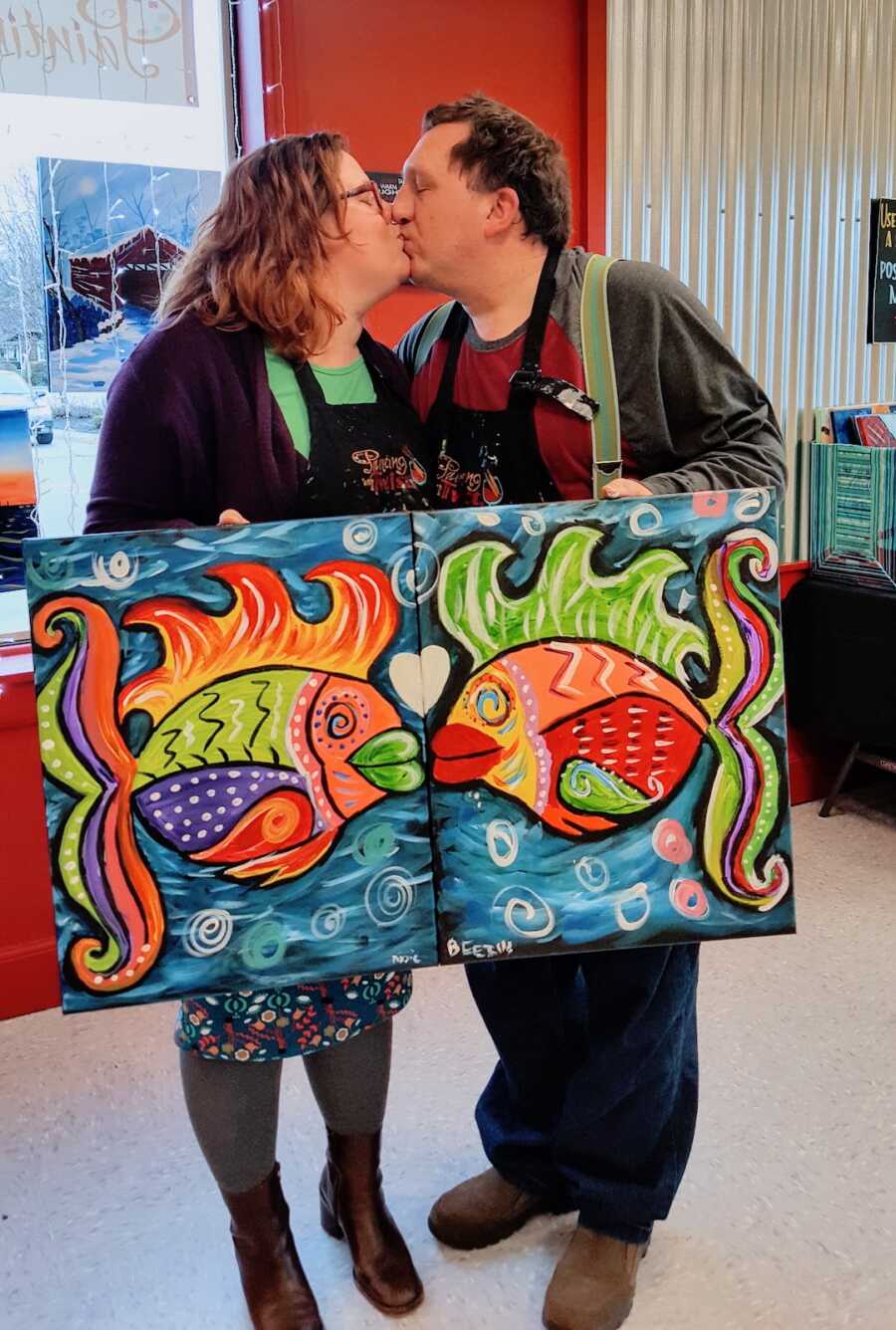 couple holds paintings they made of two fish kissing while the two of them kiss as well