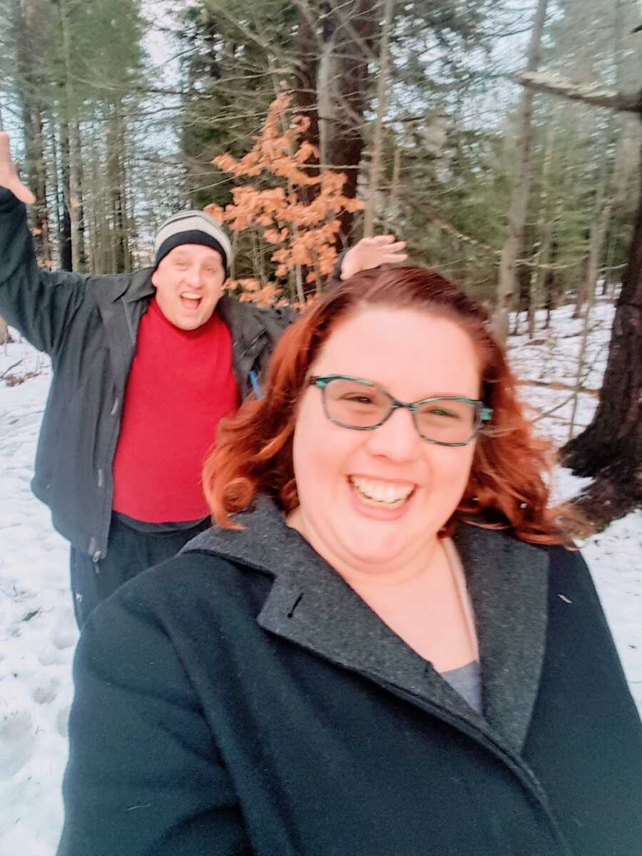 wife takes a selfie with her husband, she smiles widely while he does a silly pose behind her