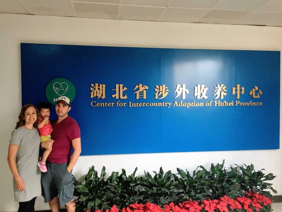 international adoptive parents hold their baby in front of an adoption agency sign