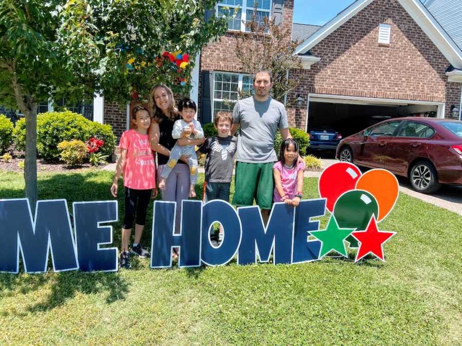 family welcomes home their adopted son with a sign in the yard, they stand with their biological and other adopted children