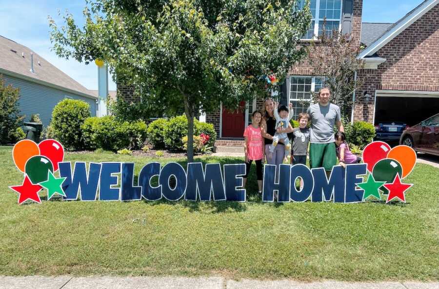 family of two biological children, two adopted children, and parents stand outside behind a welcome home sign