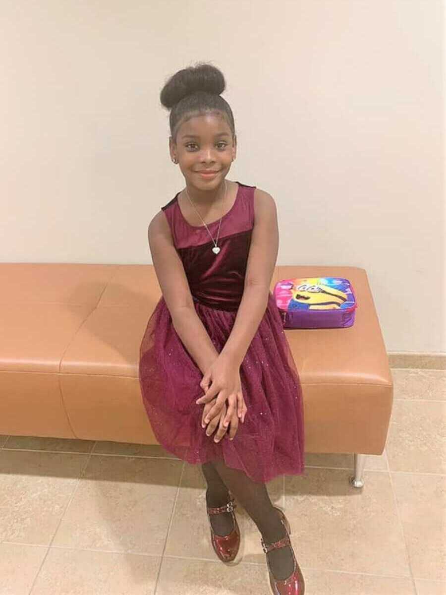 Young black girl sits in maroon dress with hands on her knee.