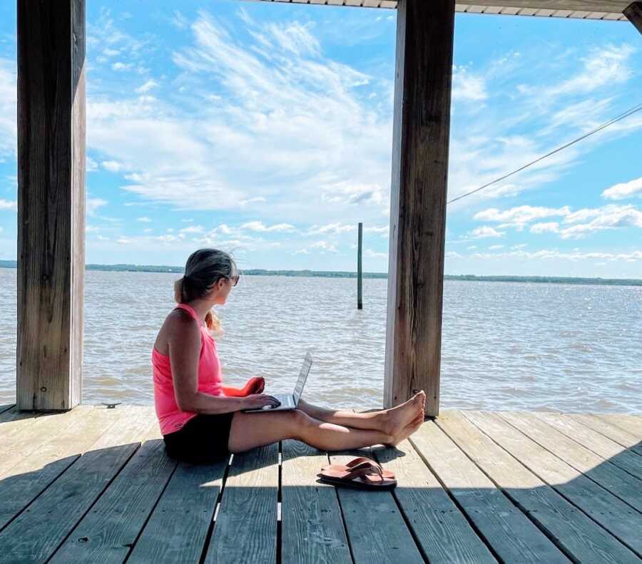 woman sits on a dock by the water with her laptop open writing a book